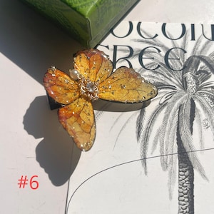 Puede incluir: Pinza para el pelo con forma de mariposa con dise&ntilde;o dorado y naranja. La pinza tiene una perla y un peque&ntilde;o diamante en el centro. El n&uacute;mero "#6" est&aacute; en rojo en la esquina inferior izquierda.