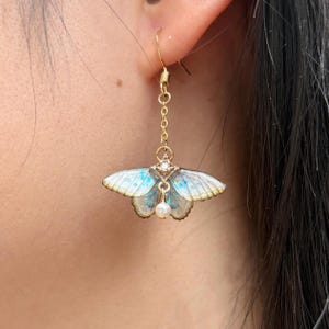 May include: A gold-colored earring featuring a butterfly design with white and blue wings. The butterfly has a small pearl pendant and is suspended from a chain. The earring is attached to a gold-colored hook.
