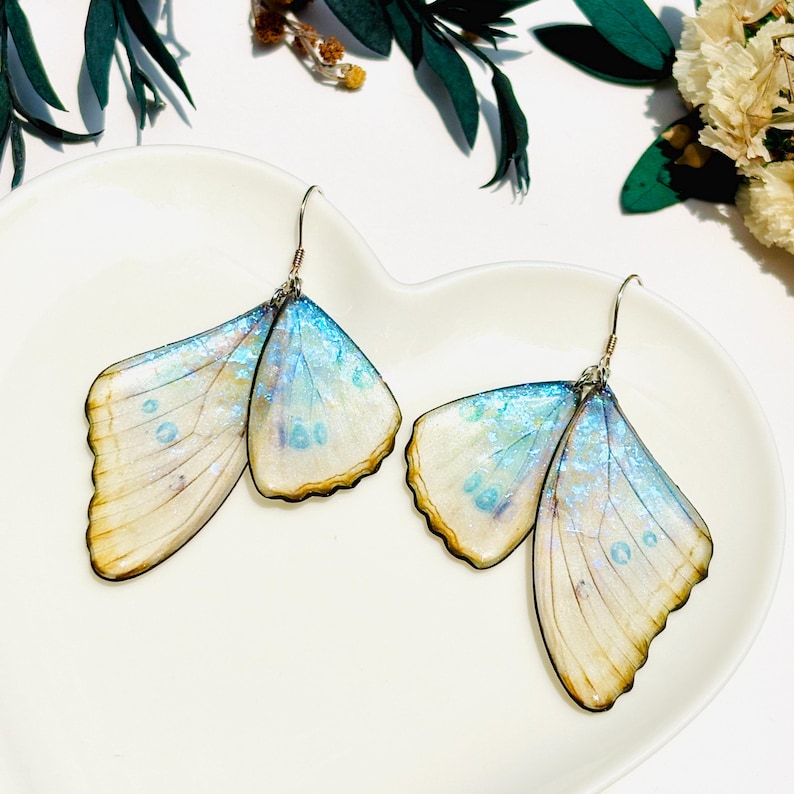Puede incluir: Un par de pendientes de mariposa con alas iridiscentes azules y blancas. Las alas est&aacute;n hechas de resina y tienen una calidad delicada y transl&uacute;cida. Los pendientes est&aacute;n unidos a ganchos de plata.