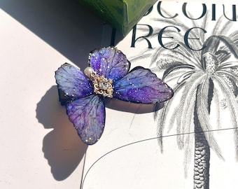 May include: A purple and blue butterfly brooch with a pearl and rhinestone embellishment. The brooch is made of resin and has a glittery finish.