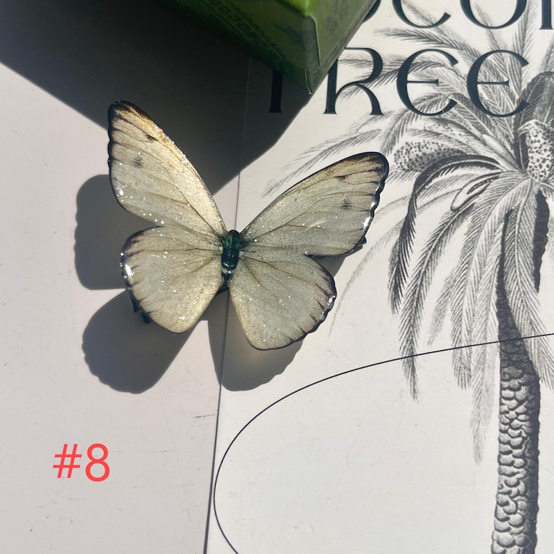 Puede incluir: Una delicada mariposa blanquecina con detalles de purpurina y bordes marrones oscuros. La mariposa est&aacute; colocada sobre una superficie blanca con una ilustraci&oacute;n de palmera y el n&uacute;mero "#8" en rojo.