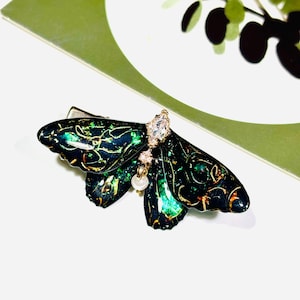 May include: A decorative hair clip in the shape of a butterfly. The clip features a dark green and gold design with a clear gemstone and a dangling pearl. The butterfly's wings have intricate patterns.