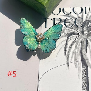 Puede incluir: Un accesorio detallado en forma de mariposa con alas verde-azuladas, acentuado con purpurina y detalles dorados. Una peque&ntilde;a gema transparente y una perla colgante adornan el centro. El n&uacute;mero "#5" es visible en la esquina inferior izquierda.