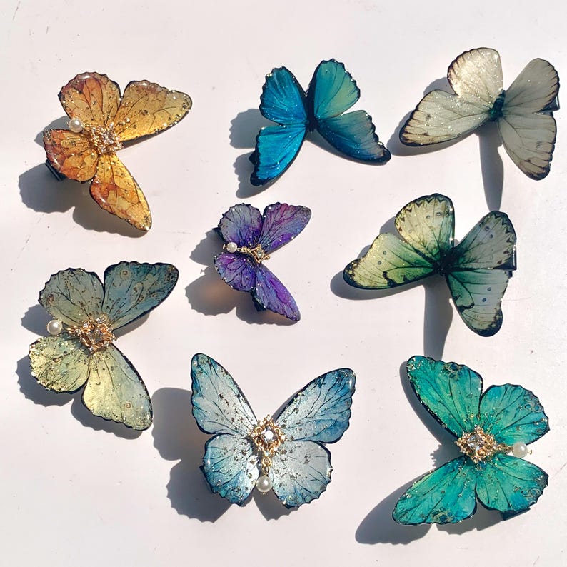 Puede incluir: Una colecci&oacute;n de pinzas para el cabello con forma de mariposa en varios colores, incluyendo dorado, azul, morado y verde. Cada pinza presenta detalles intrincados y un acento de perla, ideal para cualquier peinado.