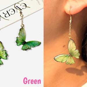 K&ouml;nnte beinhalten: Gr&uuml;ne Schmetterlingsohrringe mit goldfarbenen Ketten und Haken. Die Schmetterlingsfl&uuml;gel haben einen Farbverlauf aus Gr&uuml;nt&ouml;nen, wobei dunklere Farbt&ouml;ne die R&auml;nder umranden. Das Wort "Green" steht in rosa Schrift unten.