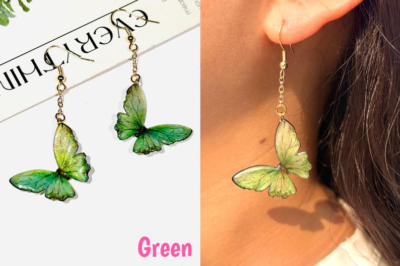 K&ouml;nnte beinhalten: Gr&uuml;ne Schmetterlingsohrringe mit goldfarbenen Ketten und Haken. Die Schmetterlingsfl&uuml;gel haben einen Farbverlauf aus Gr&uuml;nt&ouml;nen, wobei dunklere Farbt&ouml;ne die R&auml;nder umranden. Das Wort "Green" steht in rosa Schrift unten.