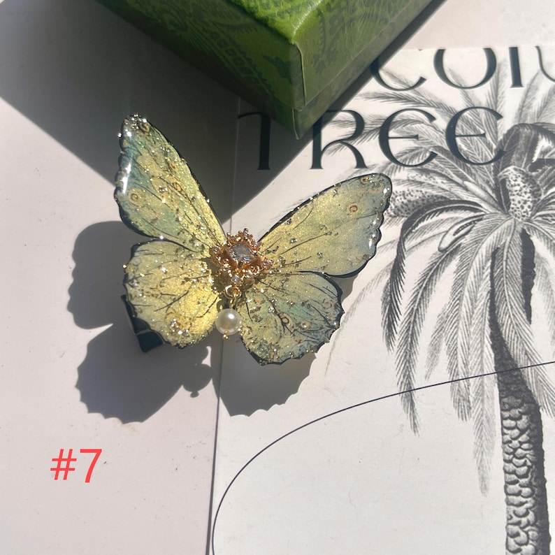 Puede incluir: Una pinza para el pelo con forma de mariposa con alas amarillas y azules, adornada con purpurina dorada y un colgante de perla. La pinza tiene un centro dorado con una gema transparente. El n&uacute;mero "#7" es visible en la esquina inferior izquierda.
