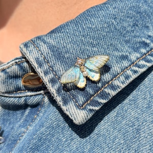Può includere: Delicata spilla a forma di farfalla con accenti azzurri e oro. Le ali presentano una sfumatura di colori, e il corpo è adornato da una piccola gemma e una perla. La spilla è appuntata su una giacca di jeans.