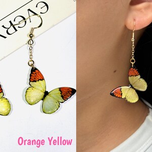 K&ouml;nnte beinhalten: Goldfarbene Schmetterlingsohrringe mit orangefarbenen und gelben Fl&uuml;geln. Die Ohrringe h&auml;ngen an einer filigranen Kette und einem Haken. Der Text "Orange Yellow" ist unten im Bild zu sehen.