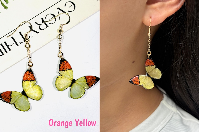 K&ouml;nnte beinhalten: Goldfarbene Schmetterlingsohrringe mit orangefarbenen und gelben Fl&uuml;geln. Die Ohrringe h&auml;ngen an einer filigranen Kette und einem Haken. Der Text "Orange Yellow" ist unten im Bild zu sehen.