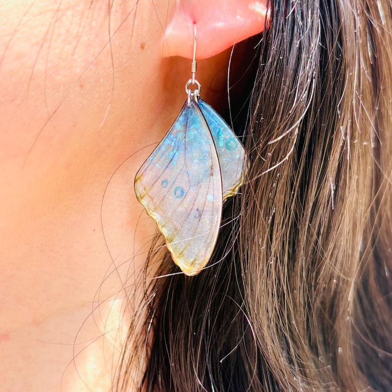 Puede incluir: Un par de pendientes de mariposa con alas iridiscentes azules y verdes. Las alas son transl&uacute;cidas y tienen un aspecto delicado y en capas.