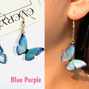 K&ouml;nnte beinhalten: Ohrringe mit blauen und lila Schmetterlingsanh&auml;ngern. Die Schmetterlingsfl&uuml;gel haben einen Farbverlauf, der von Blau nach Lila &uuml;bergeht. Die Ohrringe haben goldfarbene Haken und Ketten.