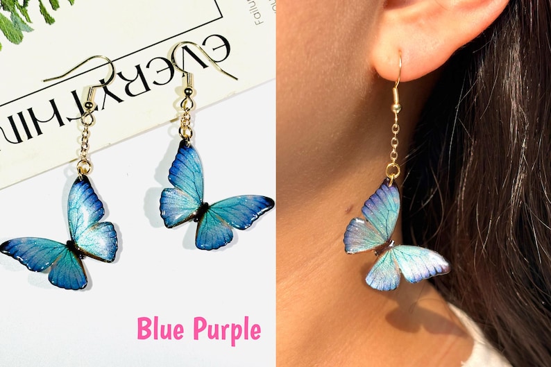 K&ouml;nnte beinhalten: Ohrringe mit blauen und lila Schmetterlingsanh&auml;ngern. Die Schmetterlingsfl&uuml;gel haben einen Farbverlauf, der von Blau nach Lila &uuml;bergeht. Die Ohrringe haben goldfarbene Haken und Ketten.
