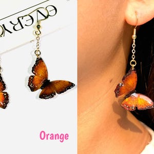 K&ouml;nnte beinhalten: Ein Paar orangefarbene Schmetterlingsohrringe mit goldfarbenen Haken und Kettenakzenten. Die Schmetterlingsfl&uuml;gel haben detaillierte Muster und dunkle R&auml;nder. Das Wort "Orange" ist in Pink geschrieben.