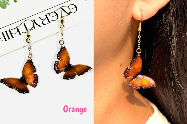 K&ouml;nnte beinhalten: Ein Paar orangefarbene Schmetterlingsohrringe mit goldfarbenen Haken und Kettenakzenten. Die Schmetterlingsfl&uuml;gel haben detaillierte Muster und dunkle R&auml;nder. Das Wort "Orange" ist in Pink geschrieben.