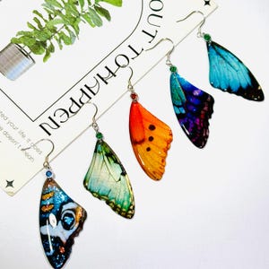 May include: Five pairs of butterfly wing earrings in various colors, including blue, green, orange, and purple. Each earring features a detailed wing design with a silver hook and a small gemstone accent. The earrings are displayed on a white surface.