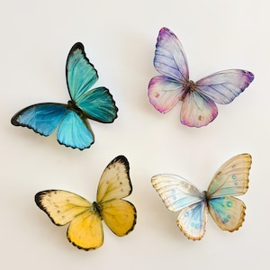 May include: Four decorative butterfly ornaments in shades of blue, purple, yellow, and white. Each butterfly has detailed wing patterns and a realistic appearance. The butterflies are arranged against a white background.