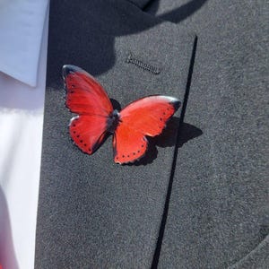 Puede incluir: Un broche de mariposa roja con detalles negros está prendido a una chaqueta de traje gris oscuro. La mariposa, con patrones detallados en las alas, se encuentra cerca de la solapa. Se ve parcialmente una camisa blanca y una corbata roja.