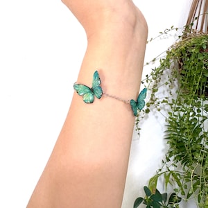 Puede incluir: Una delicada pulsera con tres dijes de mariposa turquesa. Las mariposas tienen patrones de alas detallados y están conectadas por una fina cadena plateada. La pulsera se muestra en un brazo.
