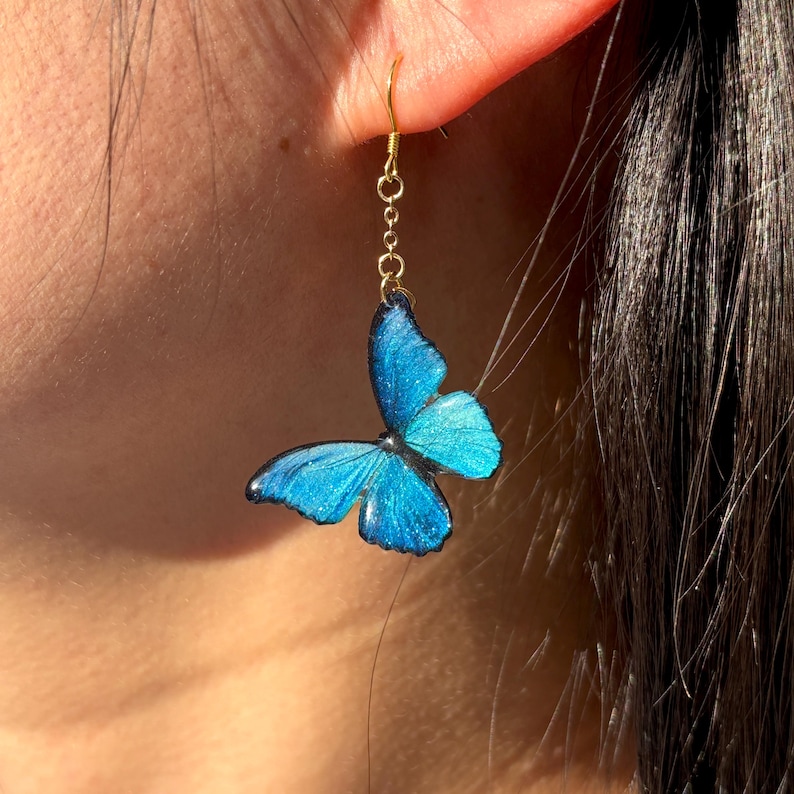K&ouml;nnte beinhalten: Goldfarbener Ohrring mit einem Anh&auml;nger in Form eines leuchtend blauen Schmetterlings. Der Schmetterling hat detaillierte Fl&uuml;gelmuster und eine dunkle Umrandung. Der Ohrring ist am Ohr einer Person befestigt.