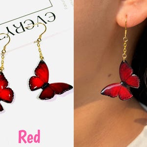 K&ouml;nnte beinhalten: Rote Schmetterlingsohrringe mit goldfarbenen Ketten und Haken. Die Schmetterlingsfl&uuml;gel sind leuchtend rot mit dunkleren Schattierungen. Die Ohrringe werden auf wei&szlig;em Hintergrund gezeigt und getragen.