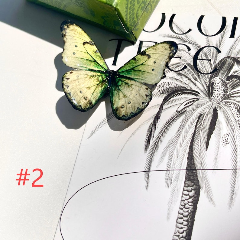 Puede incluir: Una mariposa decorativa con alas crema y verdes, detallada con manchas oscuras y un cuerpo negro. La mariposa se encuentra sobre una ilustraci&oacute;n en blanco y negro con el texto "#2" en rojo.