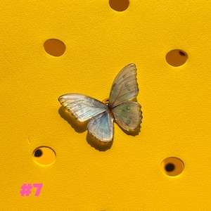 May include: A delicate butterfly-shaped charm with iridescent wings in shades of blue, green, and gold. The charm is set against a bright yellow background with circular cutouts. The number "#7" is printed in pink in the lower left corner.