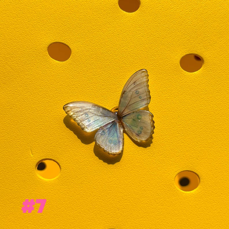 May include: A delicate butterfly-shaped charm with iridescent wings in shades of blue, green, and gold. The charm is set against a bright yellow background with circular cutouts. The number "#7" is printed in pink in the lower left corner.