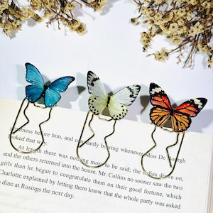 May include: Three gold-toned metal paper clips, each topped with a realistic butterfly figure. The butterflies feature distinct color patterns: one bright blue, one white with black spots, and one orange, black, and white monarch design.