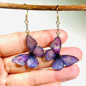 May include: A pair of butterfly-shaped earrings. The earrings feature translucent purple wings with iridescent glitter accents. They are suspended from gold-colored ear wires and chains, creating a delicate and whimsical accessory.
