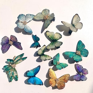 Puede incluir: Una colecci&oacute;n de pinzas para el cabello con forma de mariposa en varios colores, incluyendo azul, verde, morado y amarillo. Cada pinza presenta detalles intrincados y est&aacute; adornada con peque&ntilde;os adornos. Las pinzas est&aacute;n dispuestas sobre una superficie blanca.