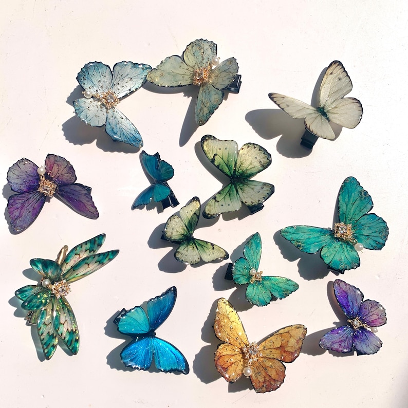 Puede incluir: Una colecci&oacute;n de pinzas para el cabello con forma de mariposa en varios colores, incluyendo azul, verde, morado y amarillo. Cada pinza presenta detalles intrincados y est&aacute; adornada con peque&ntilde;os adornos. Las pinzas est&aacute;n dispuestas sobre una superficie blanca.
