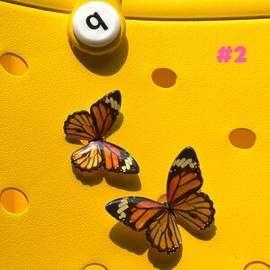 May include: Two butterfly charms with orange, yellow, and black wings are displayed on a bright yellow surface. The charms are detailed with intricate wing patterns. The number "#2" is visible in pink. A white button with a black "a" is also visible.