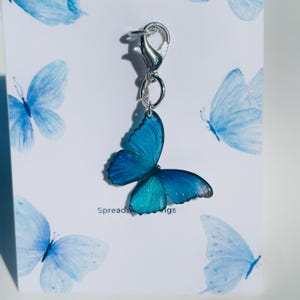 Może przedstawiać: Srebrny charm z motywem niebieskiego motyla. Motyl ma błyszczące wykończenie i jest przymocowany do srebrnego karabińczyka. Charm jest prezentowany na białej kartce z tekstem "Spread Your Wings".