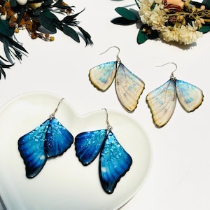 Puede incluir: Dos pares de pendientes de mariposa. Un par es azul con brillo iridiscente, el otro es blanco con brillo iridiscente.