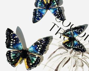 Puede incluir: Tres broches con forma de mariposa con detalles intrincados. Los broches presentan un degradado de tonos azules, verdes y amarillos, acentuados con puntos blancos. Las mariposas descansan sobre una superficie blanca con la palabra "TROPIC".