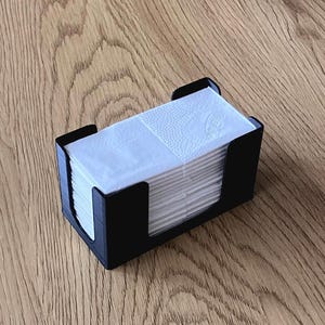 Könnte beinhalten: Ein schwarzer Taschentuchhalter mit einem Stapel weißer Taschentücher. Der Halter hat eine rechteckige Form mit einem schlichten, modernen Design. Die Taschentücher sind ordentlich gestapelt und einsatzbereit. Der Halter besteht aus einem mattschwarzen Material.
