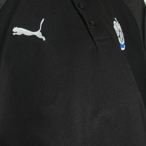 May include: Black polo shirt with a white Puma logo on the left chest. The shirt has a three-button placket and a team logo on the right chest. The fabric appears to be a textured knit.