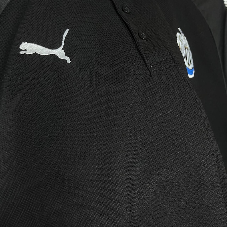 May include: Black polo shirt with a white Puma logo on the left chest. The shirt has a three-button placket and a team logo on the right chest. The fabric appears to be a textured knit.