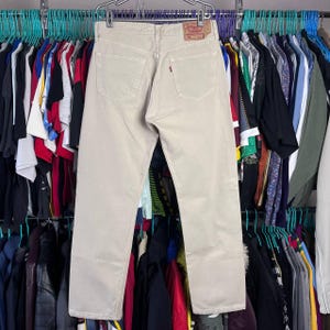 May include: Beige denim jeans with a classic five-pocket design. The jeans feature a red Levi's tag on the back pocket and are hanging on a rack with other clothing items. The pants have a straight leg cut.