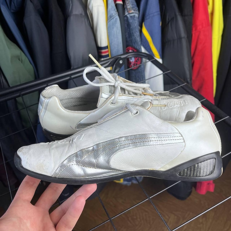 May include: White and silver Puma sneakers with a low profile design. The shoes have a leather upper with silver accents and a black sole. The laces are white, and the shoes appear to be pre-owned.