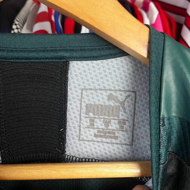 K&ouml;nnte beinhalten: Ein dunkelgr&uuml;nes und schwarzes Sportshirt mit einem grauen Innenetikett am Kragen, das das Puma-Logo und Text zeigt. Das Etikett weist die Gr&ouml;&szlig;e XS f&uuml;r UK, CAN und EUR aus, mit dem darunter gedruckten Text Made in China und Fabriqu&eacute; en Chine.
