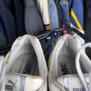 May include: Close-up of a pair of white Puma sneakers. The shoes have a silver stripe and the Puma logo on the side. The interior of the shoe is gray with the Puma logo visible. The laces are white.