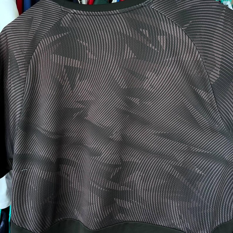 Puede incluir: Una camiseta negra de manga corta con un patr&oacute;n abstracto gris oscuro de l&iacute;neas diagonales, onduladas y entrecruzadas. La prenda tiene un cuello redondo negro s&oacute;lido y una banda horizontal negra s&oacute;lida a lo largo del dobladillo inferior.