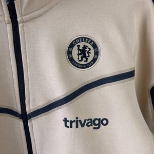 Puede incluir: Chaqueta con cremallera color crema con detalles azul marino y el logo del Chelsea Football Club. La chaqueta presenta el logotipo de Trivago y un dise&ntilde;o de rayas azul marino.