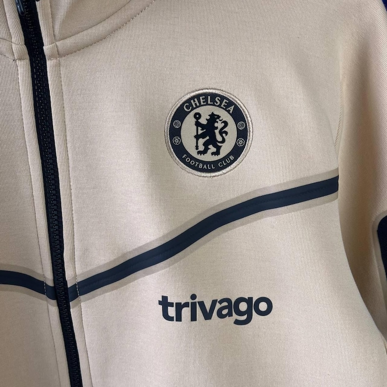 Puede incluir: Chaqueta con cremallera color crema con detalles azul marino y el logo del Chelsea Football Club. La chaqueta presenta el logotipo de Trivago y un dise&ntilde;o de rayas azul marino.