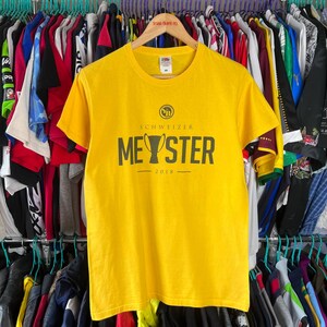 Puede incluir: Una camiseta amarilla con cuello redondo y mangas cortas. La camiseta presenta el texto "SCHWEIZER MEYSTER 2018" en verde oscuro. La camiseta est&aacute; colgada en una percha de madera entre ropa.