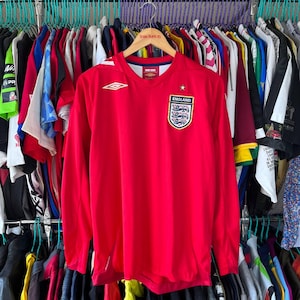 Könnte beinhalten: Ein rotes England-Fußballtrikot mit langen Ärmeln, das das England-Emblem und einen Stern zeigt. Das Umbro-Logo ist sichtbar. Das Trikot hängt an einer Kleiderstange mit anderen Kleidungsstücken.