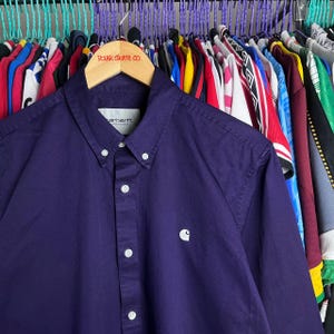 May include: A dark purple button-down shirt with white buttons and a small white logo on the chest. The shirt is hanging on a wooden hanger with the text "POLAR SKATE CO.".