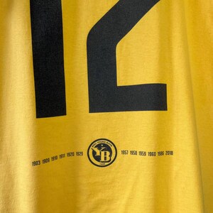 Puede incluir: Primer plano de una camiseta amarilla con un gran n&uacute;mero 12 negro. Debajo del n&uacute;mero hay un logotipo circular con el texto "BSC YOUNG BOYS" y los a&ntilde;os 1903, 1909, 1910, 1911, 1920, 1929, 1957, 1958, 1959, 1960, 1986 y 2018.
